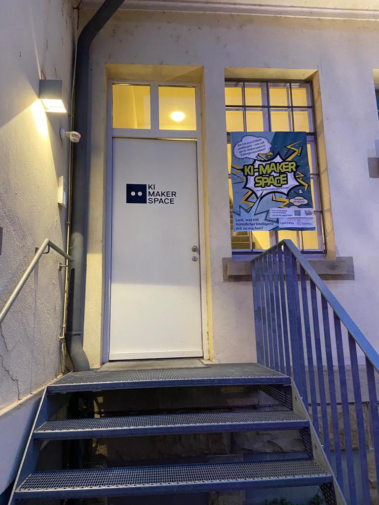 Eine weisse Tür, mit kleinen Festern über dem Rahmen, die zum KI-Makerspace führt. Die Tür ist geöffnet. Daneben eine großes Fester mit einen bunten Plakat, das für den Makerspace wirb. Zitate "Lust etwas mit KI zumachen" "Ein Ort zum Tüfteln und Basteln". Alle Fenster sind beleuchtet. 